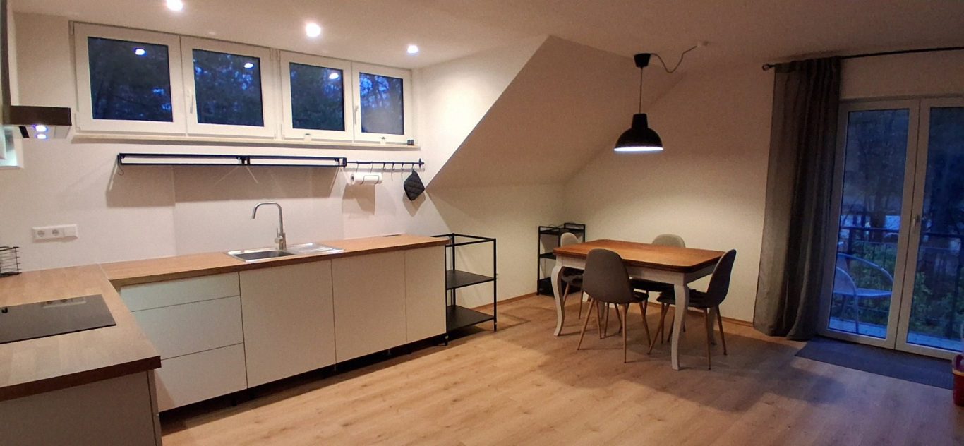 Wohnküche mit Zugang zum Balkon Moderne Küche mit Holzoberflächen, Esstisch und Fenster mit Blick ins Freie.